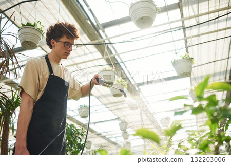 Conception of watering. Young man with curly hair and in glasses is in greenhouse Conception of watering. Young man with curly hair and in glasses is in greenhouse 132203006