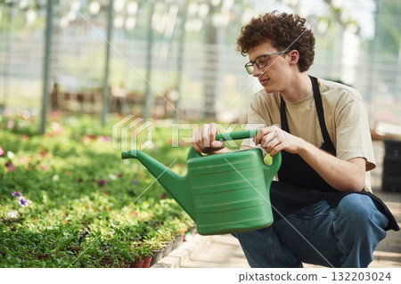 Green watering can in hands. Young man with curly hair and in glasses is in greenhouse Green watering can in hands. Young man with curly hair and in glasses is in greenhouse 132203024