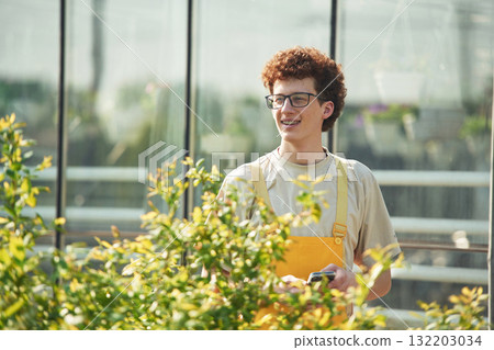 Smiling, looking forward. Young man with curly hair and in glasses is in greenhouse Smiling, looking forward. Young man with curly hair and in glasses is in greenhouse 132203034