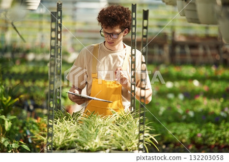Notepad in hands. Young man with curly hair and in glasses is in greenhouse Notepad in hands. Young man with curly hair and in glasses is in greenhouse 132203058