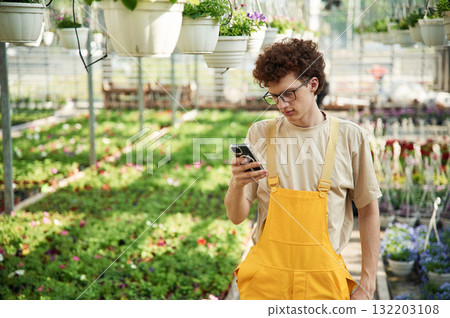 Smartphone in hands. Young man with curly hair and in glasses is in greenhouse 132203108