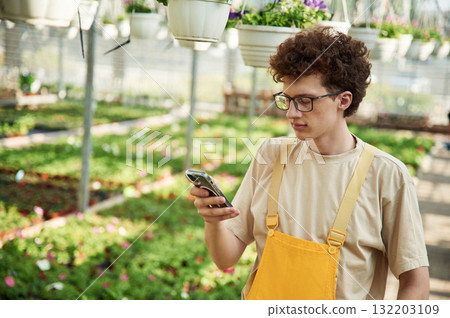 Standing, using smartphone. Young man with curly hair and in glasses is in greenhouse Standing, using smartphone. Young man with curly hair and in glasses is in greenhouse 132203109