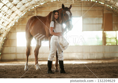 Holding cowboy hat in hand. Beautiful young woman is with horse indoors 132204629