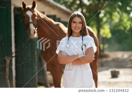 Arms crossed, standing and smiling. Female vet is with horse outdoors at the farm at daytime Arms crossed, standing and smiling. Female vet is with horse outdoors at the farm at daytime 132204832