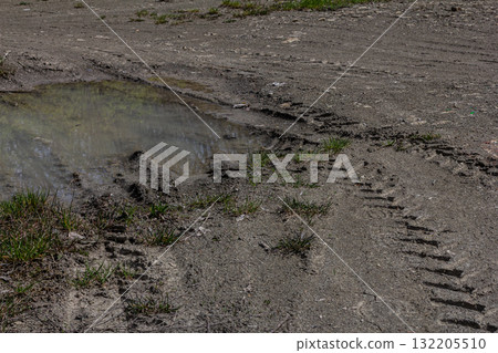 Tire tracks leading to a puddle on a dirt road surrounded by sparse grass in a rural area during daylight hours 132205510