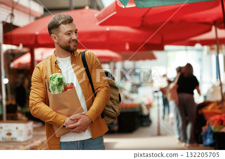 Cheerful, with products in paper bag. Handsome man is on the street market or bazaar Cheerful, with products in paper bag. Handsome man is on the street market or bazaar 132205705
