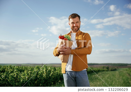Paper bag with cabbage and pepper. Handsome man is on the agricultural field at daytime Paper bag with cabbage and pepper. Handsome man is on the agricultural field at daytime 132206029