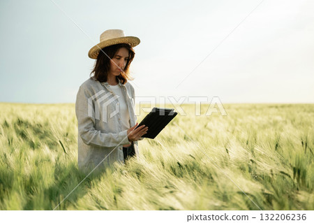 In straw hat, with tablet. Woman in white is on the agricultural wheat field In straw hat, with tablet. Woman in white is on the agricultural wheat field 132206236