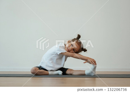 Stretching the leg. Cute little girl is sitting on the yoga mat and doing exercises Stretching the leg. Cute little girl is sitting on the yoga mat and doing exercises 132206736