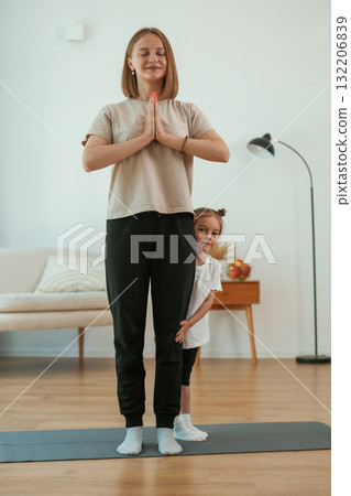 Kid is hiding behind mother's legs. Young woman with little girl are doing yoga at home Kid is hiding behind mother's legs. Young woman with little girl are doing yoga at home 132206839