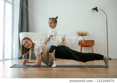 Kid is sitting on a back of woman. Young mother with little girl are doing yoga at home Kid is sitting on a back of woman. Young mother with little girl are doing yoga at home 132206870