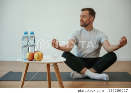 Bottles of water and apples. Yoga man is sitting on a mat at home Bottles of water and apples. Yoga man is sitting on a mat at home 132206927