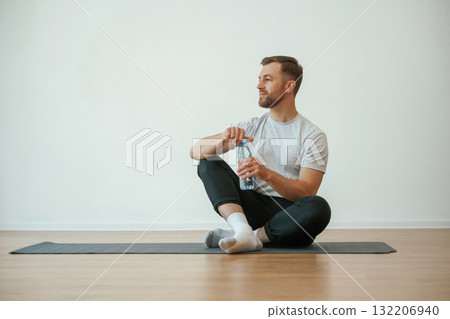 Taking a break, with bottle of water. Yoga man is sitting on a mat at home Taking a break, with bottle of water. Yoga man is sitting on a mat at home 132206940