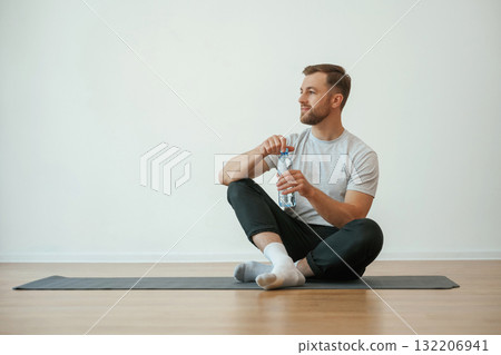 Taking a break, with bottle of water. Yoga man is sitting on a mat at home Taking a break, with bottle of water. Yoga man is sitting on a mat at home 132206941