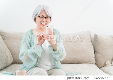 An elderly woman sitting on a sofa and using a smartphone (smile, fun) 132207448