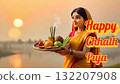 Smiling woman in red and yellow saree performing Chhath Puja, offering fruits and incense to the sun god at sunset over a calm water body. 132207908