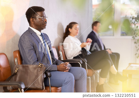 Serious-minded African male candidate siting on chair and waiting for job interview in hallway Serious-minded African male candidate siting on chair and waiting for job interview in hallway 132210551