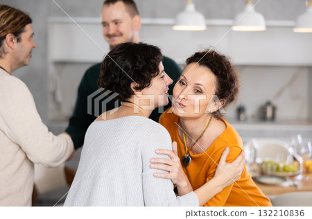 Women hug each other and kiss on the cheek while greeting each other before a festive dinner. Men shaking hands in background Women hug each other and kiss on the cheek while greeting each other before a festive dinner. Men shaking hands in background 132210836