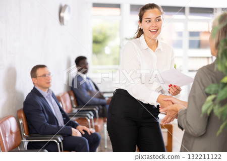 Smiling young girl makes deal, shaking hands with elderly woman colleague while standing in office. 132211122