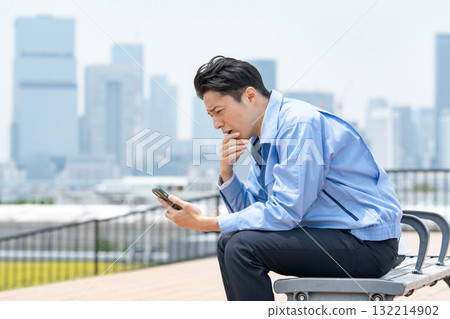 一名身穿工作服的男子正看著他的智慧型手機,憂心忡忡 一名身穿工作服的男子正看著他的智慧型手機,憂心忡忡 132214902