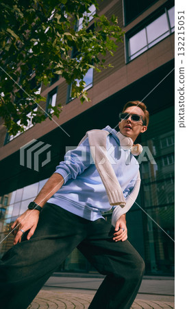 Man in blue hoodie with headphones walking near glass building and trees 132215051
