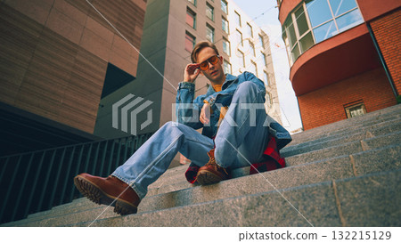 Young man sitting on stairs wearing denim outfit and orange glasses 132215129