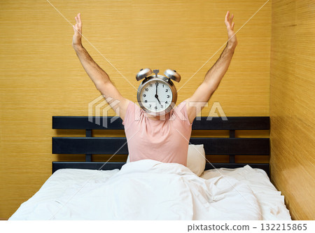 Happy man with alarm clock instead of head stretching on bed at home 132215865