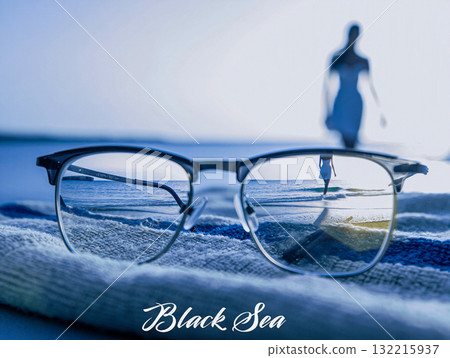 Black Sea reflections through eyeglasses. Glasses on a towel reflect the serene waters of the Black Sea. In the blurred background, a woman in a dress walks along the shore. Black Sea reflections through eyeglasses. Glasses on a towel reflect the serene waters of the Black Sea. In the blurred background, a woman in a dress walks along the shore. 132215937