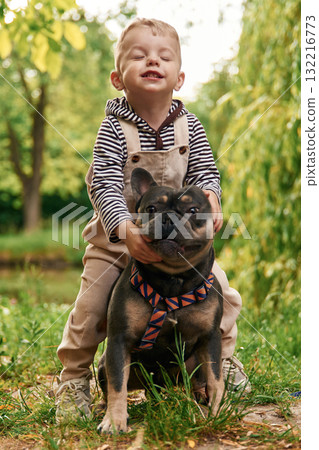 Riding the animal, sitting on it. Cute little boy is with French Bulldog outdoors in the park 132216773