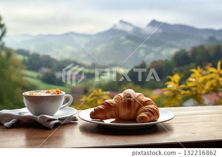 a cup of coffee and a fresh croissant on the table with a view of a beautiful landscape, still life, mountains and forest, haze at dawn 132216862