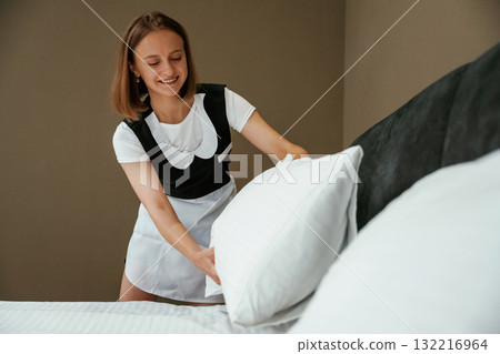 In uniform, taking care of bed. Young chambermaid is in hotel room In uniform, taking care of bed. Young chambermaid is in hotel room 132216964