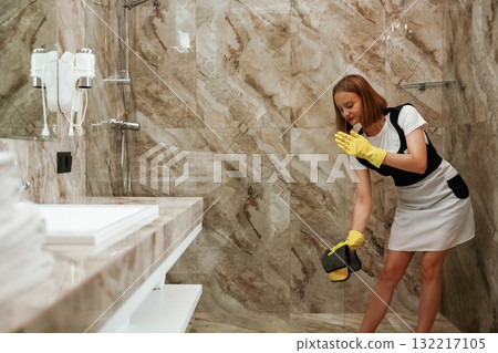 Glass in the bathroom is getting cleaned. Young chambermaid is in hotel room Glass in the bathroom is getting cleaned. Young chambermaid is in hotel room 132217105