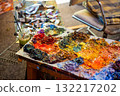 Artist creative workspace with oil paint palette, colorful brushstrokes, paint tubes, glass jar, chair and canvas in the background. Real studio atmosphere full of artistic energy. 132217202