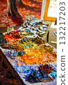 Colorful oil paint palette with messy brushstrokes, vibrant pigments, scattered tubes and a finished canvas in the background. Artistic chaos as a symbol of creativity and inspiration 132217203