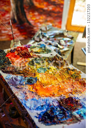 Colorful oil paint palette with messy brushstrokes, vibrant pigments, scattered tubes and a finished canvas in the background. Artistic chaos as a symbol of creativity and inspiration Colorful oil paint palette with messy brushstrokes, vibrant pigments, scattered tubes and a finished canvas in the background. Artistic chaos as a symbol of creativity and inspiration 132217203