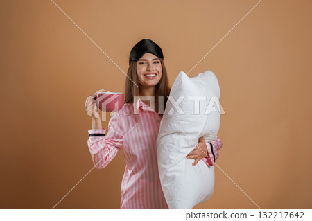 With cup of drink, in pajama and with pillow. Young pretty woman is against background in the studio 132217642