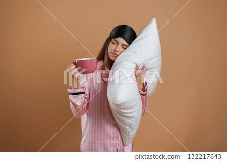 With cup of drink, in pajama and with pillow. Young pretty woman is against background in the studio With cup of drink, in pajama and with pillow. Young pretty woman is against background in the studio 132217643