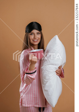 With cup of drink, in pajama and with pillow. Young pretty woman is against background in the studio With cup of drink, in pajama and with pillow. Young pretty woman is against background in the studio 132217644
