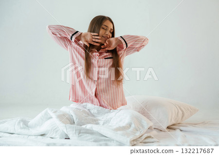 Front view, in pink pajama, waking up. Young pretty woman is against background in the studio 132217687