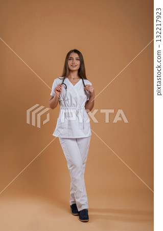 In doctor coat, with stethoscope. Young pretty woman is against background in the studio In doctor coat, with stethoscope. Young pretty woman is against background in the studio 132217923