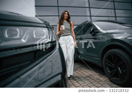 Holding cup of coffee. Attractive young woman is outdoors near her brand new car Holding cup of coffee. Attractive young woman is outdoors near her brand new car 132218332