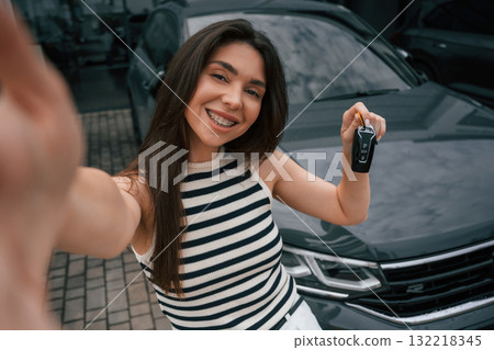 Making selfie, holding keys, happy owner. Attractive young woman is outdoors near her brand new car 132218345