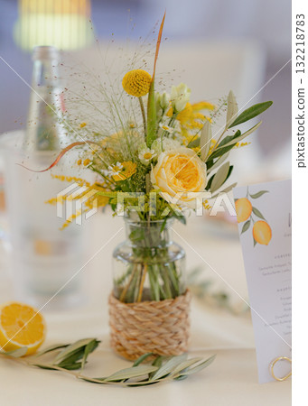 Floral arrangement with yellow and white flowers in a glass jar on a wedding table. Fresh lemon half, greenery, and menu card with a lemon design create a charming, rustic atmosphere Floral arrangement with yellow and white flowers in a glass jar on a wedding table. Fresh lemon half, greenery, and menu card with a lemon design create a charming, rustic atmosphere 132218783