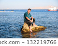 A blond man in clothes sits on a rock in the sea far from the shore 132219766