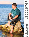 A blond man in clothes sits on a rock in the sea far from the shore 132219767