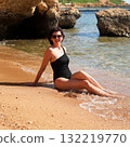 A brunette woman in a one-piece black bathing suit sits on the sand overlooking the rocks and the sea 132219770