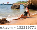 A brunette woman in a one-piece black bathing suit sits on the sand overlooking the rocks and the sea 132219771
