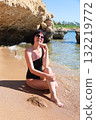 A brunette woman with glasses in a black bathing suit sits on the sand overlooking the sea, with her legs tucked under her, enjoying the sun's rays 132219772