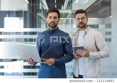 Two confident businessmen standing in an office, one holding a laptop and the other a tablet, representing teamwork, technology, and a modern corporate environment Two confident businessmen standing in an office, one holding a laptop and the other a tablet, representing teamwork, technology, and a modern corporate environment 132219891