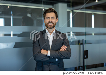 Businessman with a beard and dark suit standing confidently with arms crossed in a modern office building, looking directly at the camera with a friendly smile 132219892